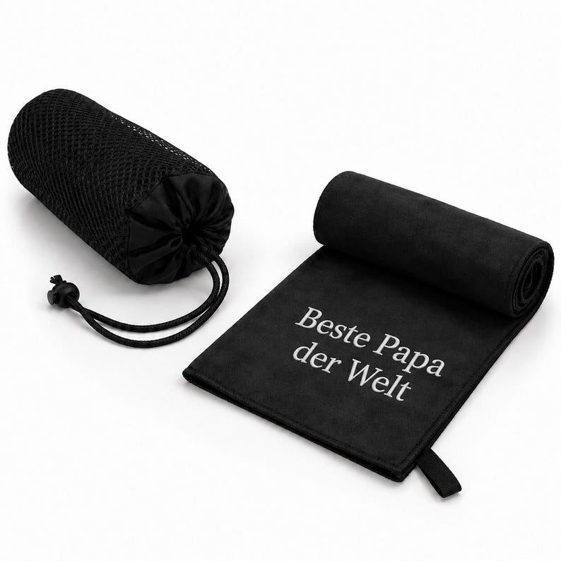 Op de afbeelding: Een zwarte microvezel handdoek met de witte tekst "Beste Papa der Welt" erop gedrukt, gedeeltelijk opgerold, vergezeld van een zwarte mesh opbergtas met trekkoordsluiting, ontworpen voor compacte draagbaarheid en opslag.