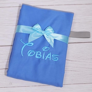 Op de afbeelding: Een felblauw rechthoekig stoffen item met de geborduurde naam Tobias in een gestileerd lettertype. Het item is omwikkeld met een lichtblauw satijnen lint dat in het midden tot een strik is geknoopt en heeft een grijze elastische band aan de zijkant.