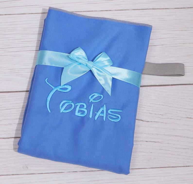 Op de afbeelding: Een felblauw rechthoekig stoffen item met de geborduurde naam Tobias in een gestileerd lettertype. Het item is omwikkeld met een lichtblauw satijnen lint dat in het midden tot een strik is geknoopt en heeft een grijze elastische band aan de zijkant.