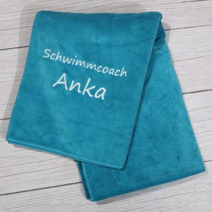 Op de afbeelding: Een gevouwen talingkleurige handdoek met wit geborduurde tekst die Schwimmcoach Anka leest. De stof heeft een zachte, pluche textuur en is netjes gelaagd, wat de levendige effen kleur en de duidelijke, gestikte letters op het vooroppervlak toont.