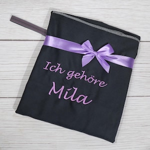 Op de afbeelding: Een zwart stoffen tasje met een paars satijnen lint dat aan de voorzijde tot een strik is geknoopt. De tekst Ich geh&ouml;re Mila is in paars garen onder het lint geborduurd. Een dun grijs elastisch bandje steekt uit de linkerbovenhoek.