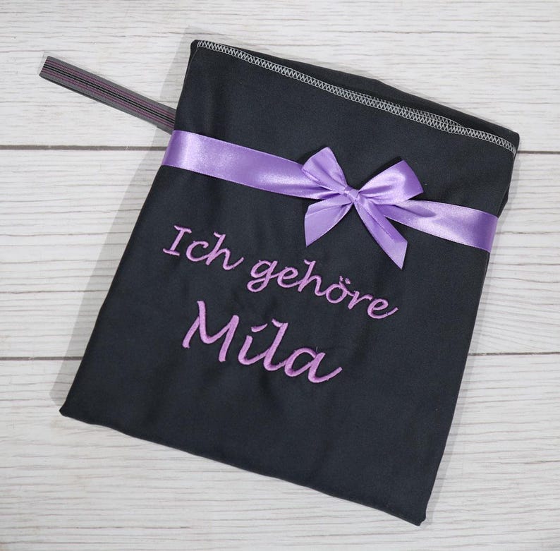 Op de afbeelding: Een zwart stoffen tasje met een paars satijnen lint dat aan de voorzijde tot een strik is geknoopt. De tekst Ich geh&ouml;re Mila is in paars garen onder het lint geborduurd. Een dun grijs elastisch bandje steekt uit de linkerbovenhoek.