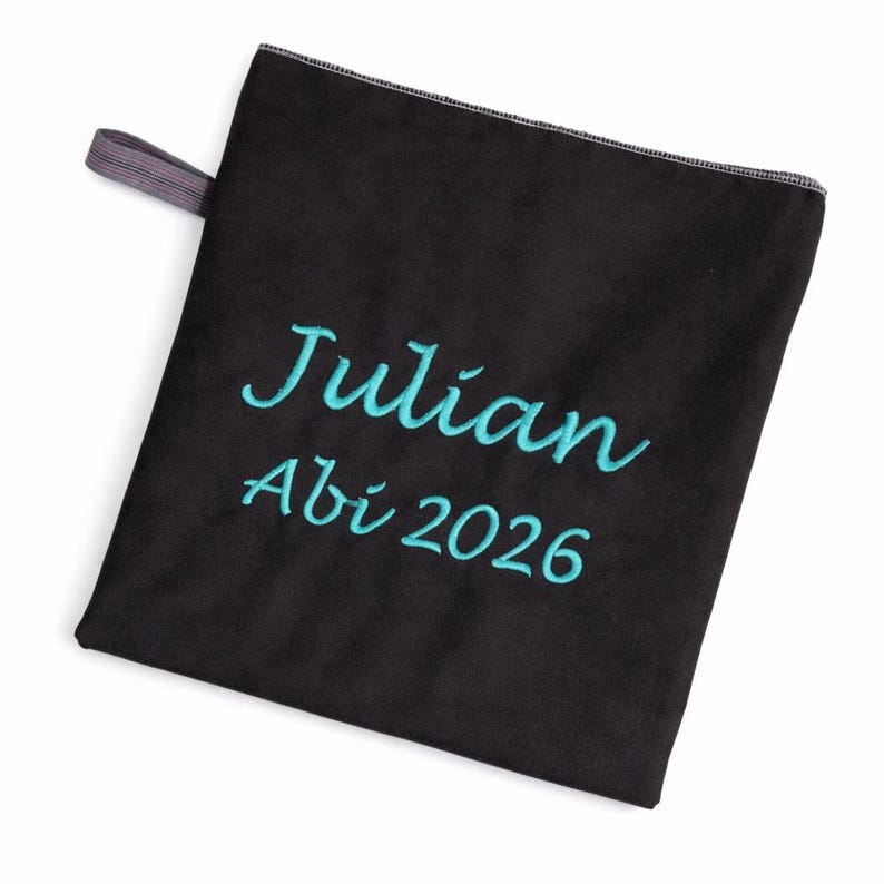 Op de afbeelding: Een vierkante zwarte stoffen doek met een kleine grijze ophanglus aan de linkerbovenhoek. De doek is geborduurd met de naam Julian en de tekst Abi 2026 in een helder turquoise cursief lettertype in het midden van de voorzijde.