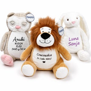 Op de afbeelding: Drie pluche knuffeldieren, waaronder een beige uil met gesloten ogen, een bruine leeuw met een pluizige manen en een wit konijn met lange oren. Elk speelgoed heeft geborduurde tekst op de witte buik, inclusief namen en geboortegegevens.