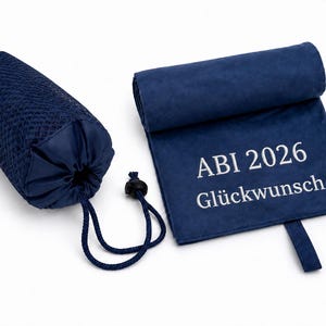 Op de afbeelding: Een marineblauwe microvezel handdoek met de witte geborduurde tekst ABI 2026 Gl&uuml;ckwunsch, vergezeld door een bijpassende marineblauwe mesh opbergtas met trekkoordsluiting, gepresenteerd tegen een effen witte achtergrond.
