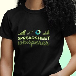 May include: Black t-shirt with the text "SPREADSHEET whisperer" in a green gradient font. The shirt features a graphic of charts and graphs. The person in the image is wearing black jeans and glasses.