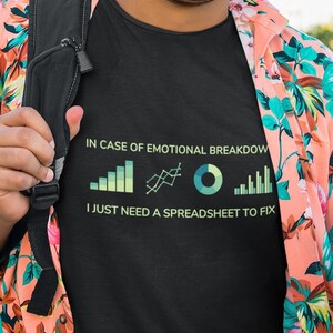 May include: A black t-shirt with the text "IN CASE OF EMOTIONAL BREAKDOWN I JUST NEED A SPREADSHEET TO FIX" in white and green. The shirt features a graphic of charts and graphs. The person is wearing a floral shirt and holding a coffee cup.