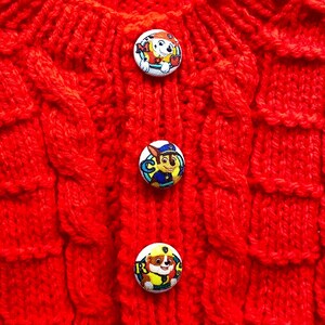May include: A close-up shot of a vibrant red knitted item, fastened with three circular buttons. The buttons display colourful cartoon characters. The knit has a textured cable pattern. The item is likely children's clothing.