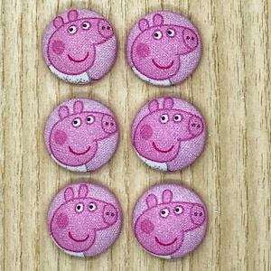 May include: A set of six round fabric-covered buttons, each featuring a printed graphic of the pink cartoon character Peppa Pig. The buttons are arranged in two vertical columns of three, displaying the character's face with a smiling expression.