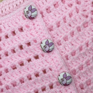 May include: A light pink, crocheted baby cardigan with three decorative buttons. The buttons feature a cartoon pig design in shades of purple and pink. The cardigan has an open weave pattern.