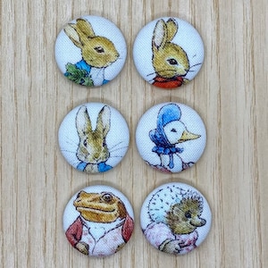 May include: Six circular fabric buttons with illustrations of characters from Beatrix Potter's stories. The buttons feature Peter Rabbit, a duck, a frog, and a hedgehog, with colourful designs on a white background.