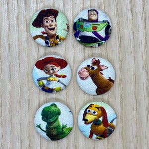 May include: A set of six circular fabric-covered buttons featuring printed illustrations of characters from the Toy Story franchise, including Woody, Buzz Lightyear, Jessie, Bullseye, Rex, and Slinky Dog, arranged in two vertical columns of three.