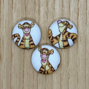 May include: Three circular fabric buttons featuring Tigger from Winnie the Pooh. Each button has a white background with a colourful image of Tigger. The buttons are orange and brown with a pink mouth and are set against a light wood grain surface.