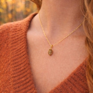 May include: A gold-coloured necklace with a small, detailed pinecone pendant. The pendant hangs from a delicate chain. The person in the image is wearing an orange jumper. The background is blurred, suggesting an outdoor setting.