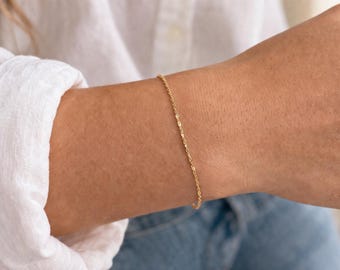 May include: A delicate gold bracelet with a chain-link design. The bracelet is worn on a wrist, and the gold color contrasts with the skin tone. The bracelet is a simple, elegant piece of jewelry.