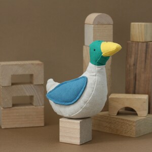 May include: A plush toy duck with a white body, blue wings, a green head, and a yellow beak. The duck is sitting on a wooden block, surrounded by other wooden blocks.
