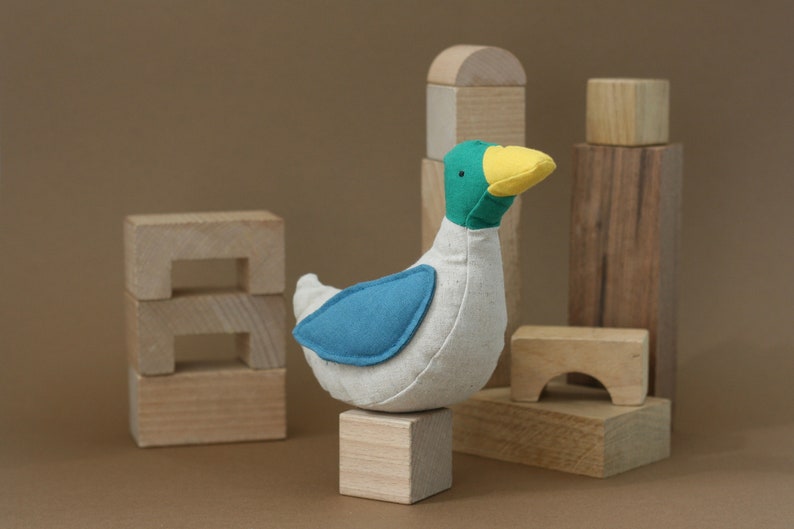 May include: A plush toy duck with a white body, blue wings, a green head, and a yellow beak. The duck is sitting on a wooden block, surrounded by other wooden blocks.
