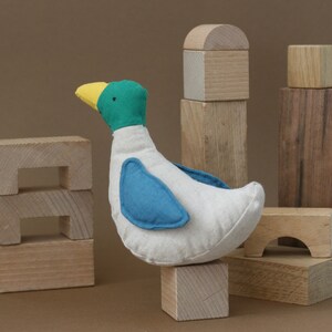 May include: A white fabric duck toy with blue wings and a yellow beak, standing on a wooden block. The duck is surrounded by wooden blocks of various sizes and shapes.