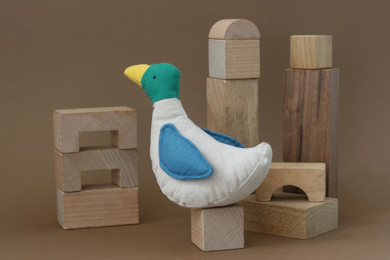 May include: A white fabric duck toy with blue wings and a yellow beak, standing on a wooden block. The duck is surrounded by wooden blocks of various sizes and shapes.