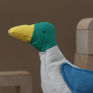 May include: A plush toy shaped like a bird with a yellow beak, green head, and blue wing patch. The bird is made of soft fabric and has a white body.