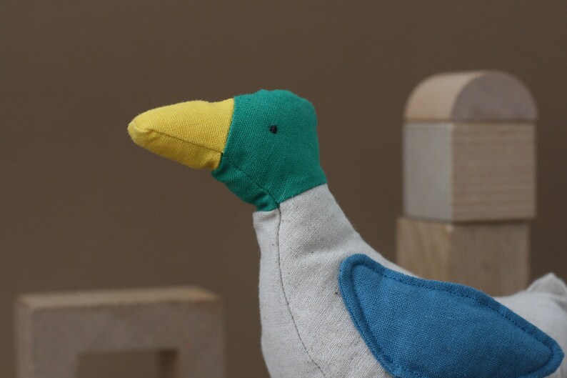 May include: A plush toy shaped like a bird with a yellow beak, green head, and blue wing patch. The bird is made of soft fabric and has a white body.