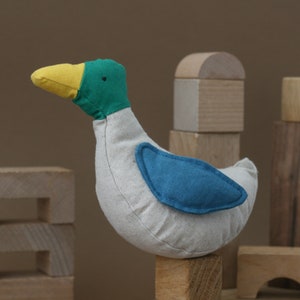 May include: A plush toy duck with a yellow beak, green head, and blue wing. The duck is sitting on a wooden block.