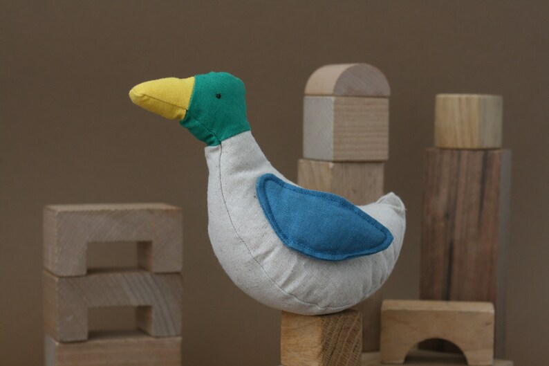 May include: A plush toy duck with a yellow beak, green head, and blue wing. The duck is sitting on a wooden block.