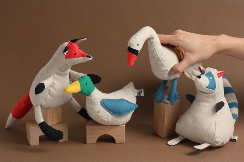 May include: Four plush stuffed animals: a fox with a red tail, a duck with a green head and blue wings, a swan with a red beak, and a raccoon with a blue face and striped tail.