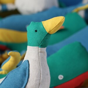 May include: A plush toy duck with a green head, yellow beak, and blue and white body. The duck is sitting on a colourful background with other plush toys.