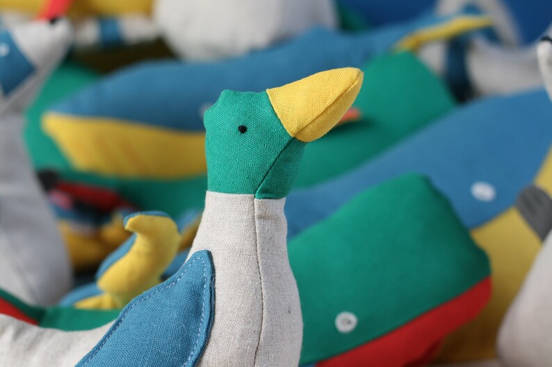 May include: A plush toy duck with a green head, yellow beak, and blue and white body. The duck is sitting on a colourful background with other plush toys.