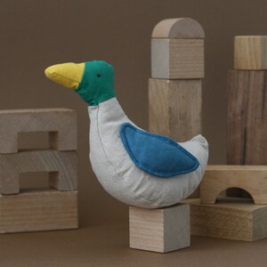 May include: A plush toy duck with a white body, yellow beak, green head, and blue wing patch. The duck is sitting on a wooden block, surrounded by other wooden blocks.