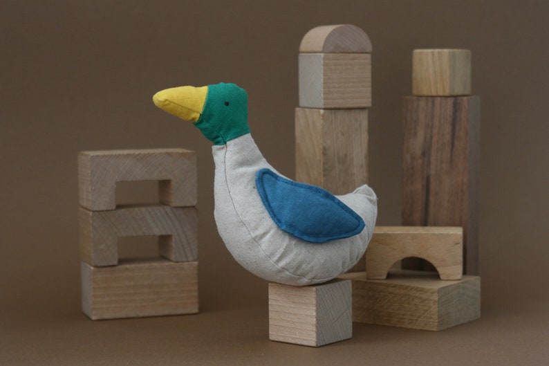May include: A plush toy duck with a white body, yellow beak, green head, and blue wing patch. The duck is sitting on a wooden block, surrounded by other wooden blocks.