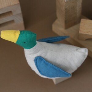 May include: A handmade plush toy duck with a yellow beak, green head, and blue wings. The duck is made of soft, white fabric and has a simple, charming design.