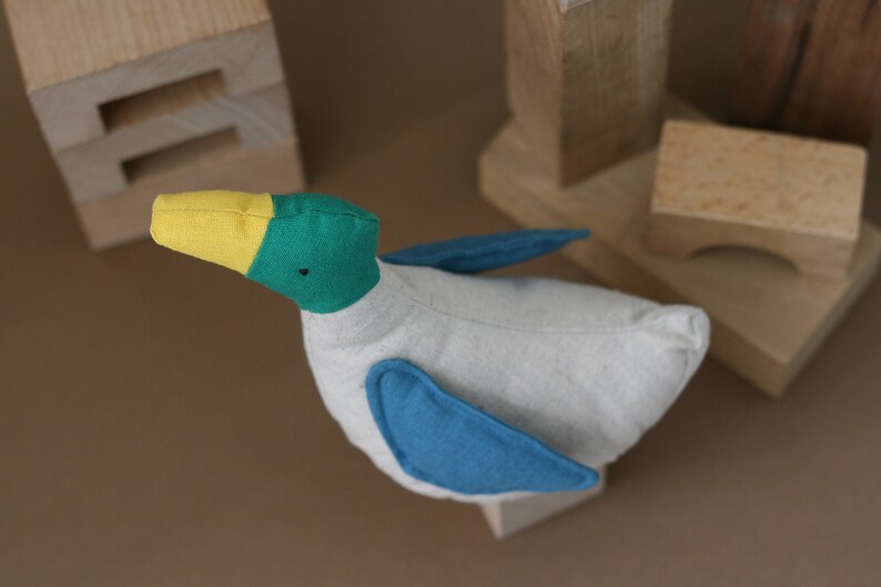 May include: A handmade plush toy duck with a yellow beak, green head, and blue wings. The duck is made of soft, white fabric and has a simple, charming design.