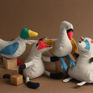 May include: A set of five plush toys, including a duck, fox, swan, raccoon, and a set of wooden blocks. The toys are made of a soft, light grey fabric with colourful accents. The duck is blue and green, the fox is red and black, the swan is red and blue, and the raccoon is blue and white.