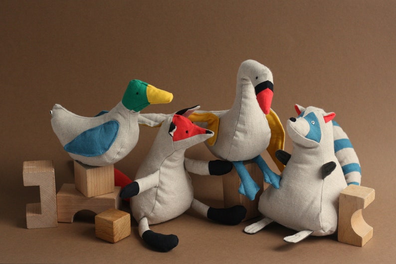 May include: A set of five plush toys, including a duck, fox, swan, raccoon, and a set of wooden blocks. The toys are made of a soft, light grey fabric with colourful accents. The duck is blue and green, the fox is red and black, the swan is red and blue, and the raccoon is blue and white.