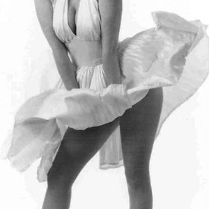 May include: A black and white photograph of a woman wearing a white dress with a flowing skirt. The dress is cinched at the waist and has a halter neckline. The woman is wearing her hair in a blonde updo and has a slight smile on her face.