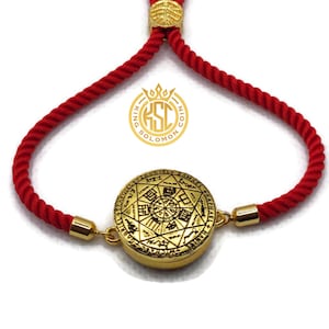 May include: Gold-colored bracelet with a circular pendant featuring intricate engravings and a red rope band. The bracelet has gold-colored accents and a sliding knot closure. The pendant has a detailed design with text and symbols.