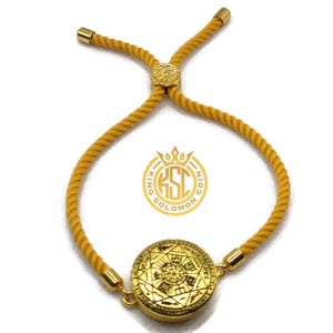 May include: Gold-colored adjustable bracelet with a circular pendant featuring an intricate design. The bracelet has a mustard-yellow rope-like band and gold-toned metal accents. The pendant has a detailed pattern and the logo "KING SOLOMON COIN".