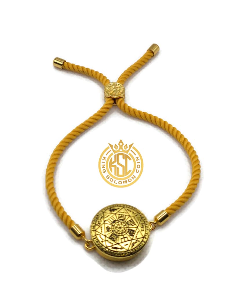 May include: Gold-colored adjustable bracelet with a circular pendant featuring an intricate design. The bracelet has a mustard-yellow rope-like band and gold-toned metal accents. The pendant has a detailed pattern and the logo "KING SOLOMON COIN".