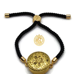 May include: Gold-colored bracelet with a circular pendant featuring an engraved design and black rope-like cord. The bracelet has gold-colored end caps and a gold-colored adjustable clasp. The pendant has the text "KING SOLOMON" on it.