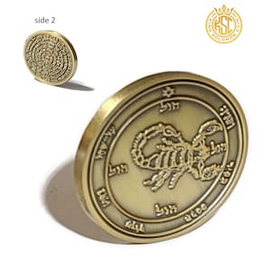 Può includere: Una moneta di metallo color oro con un designo a scorpione e testo ebraico su un lato. L'altro lato della moneta ha un design circolare con testo ebraico.