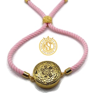 May include: A gold-colored bracelet with a pink cord. The bracelet features a circular pendant with intricate engravings and the text "KING SOLOMON COIN". The cord is adjustable with gold-colored accents.