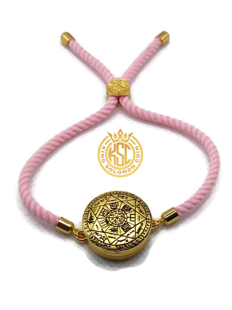 May include: A gold-colored bracelet with a pink cord. The bracelet features a circular pendant with intricate engravings and the text "KING SOLOMON COIN". The cord is adjustable with gold-colored accents.