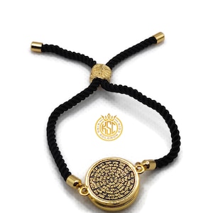 May include: A gold-colored bracelet with a black rope band. The round pendant features engraved text. The bracelet has gold-colored end caps and a gold-colored adjustable clasp. The brand logo is visible.