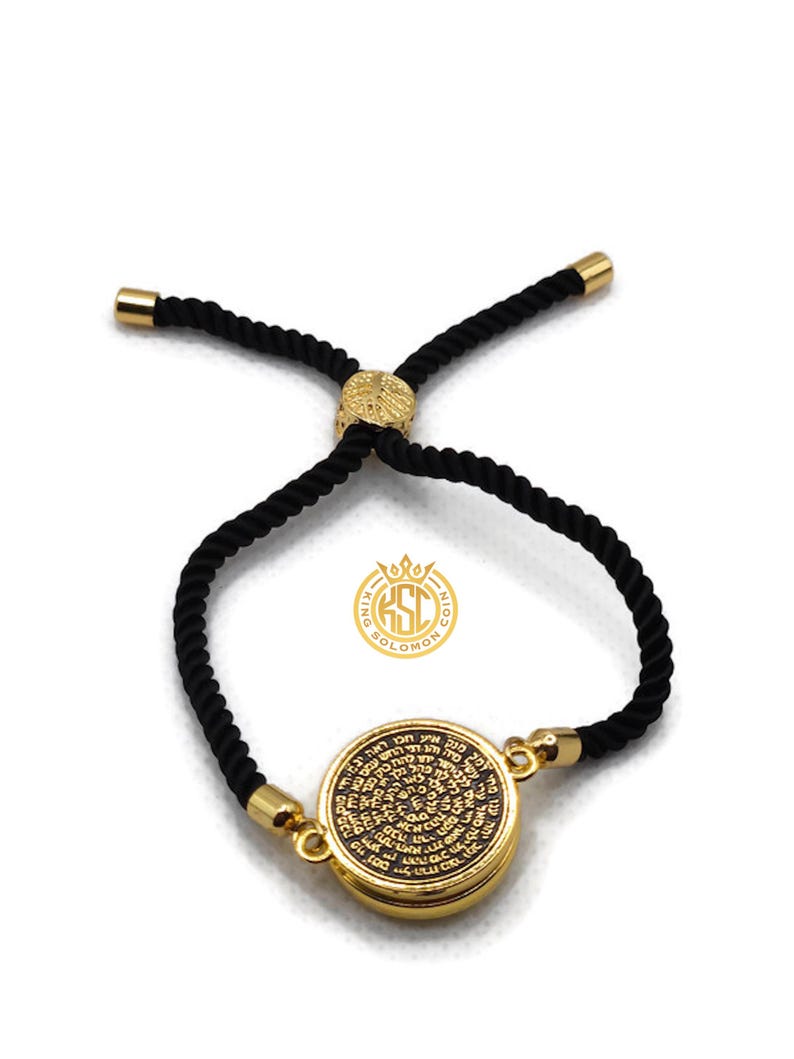 May include: A gold-colored bracelet with a black rope band. The round pendant features engraved text. The bracelet has gold-colored end caps and a gold-colored adjustable clasp. The brand logo is visible.