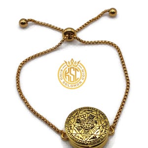 May include: Gold-toned adjustable bracelet featuring a circular pendant with intricate engraved symbols and text. The pendant is the focal point, connected to a delicate chain with a sliding bead for adjustment. The bracelet has a polished finish.