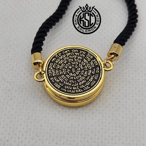 May include: A gold-colored pendant with black text and a black rope necklace. The pendant has a circular design with Hebrew text and the words "King Solomon Coin" in a logo. The necklace is displayed against a light background.