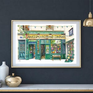 May include: Watercolor painting of the Shakespeare and Company bookstore in Paris, France. The shop front is painted in shades of green with the store's name in white lettering. The artwork is framed in a gold-colored frame.