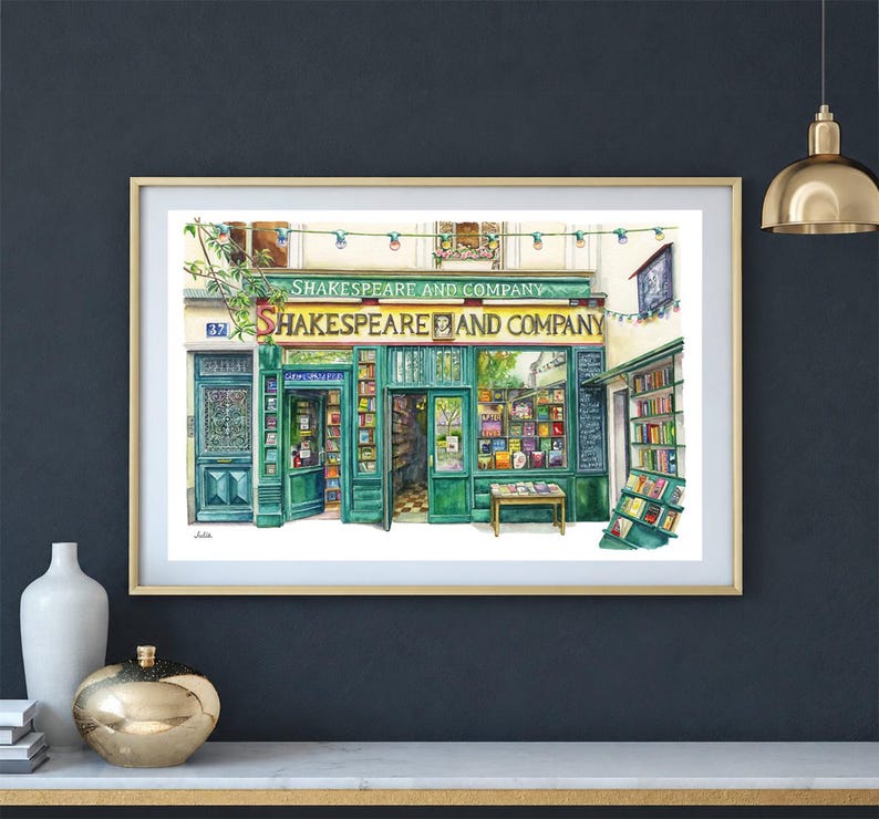 May include: Watercolor painting of the Shakespeare and Company bookstore in Paris, France. The shop front is painted in shades of green with the store's name in white lettering. The artwork is framed in a gold-colored frame.